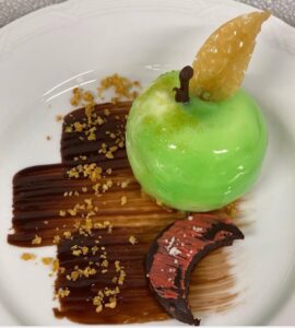 A gourmet dessert plating featuring a vibrant green apple-shaped confection atop a chocolate swirl, accompanied by a chocolate piece and garnished with a golden crisp.